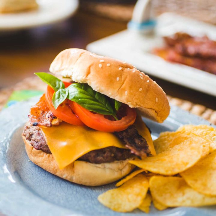 A bacon cheddar burger has layers of fresh tomato and fresh basil and sits next to a serving of potato chips.