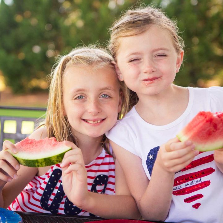 Two young girls wearing red, white, and blue tees are holding fresh slices of watermelon at a backyard BBQ party.