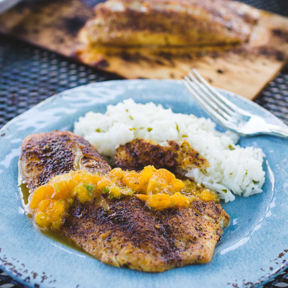 Yellow mojito salsa is drizzled over a spicy grilled tilapia on a blue plate next to a serving of white rice.