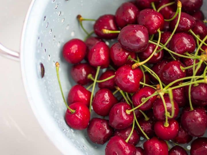 A blue strainer holds a pile of fresh cherries with stems for rinsing.