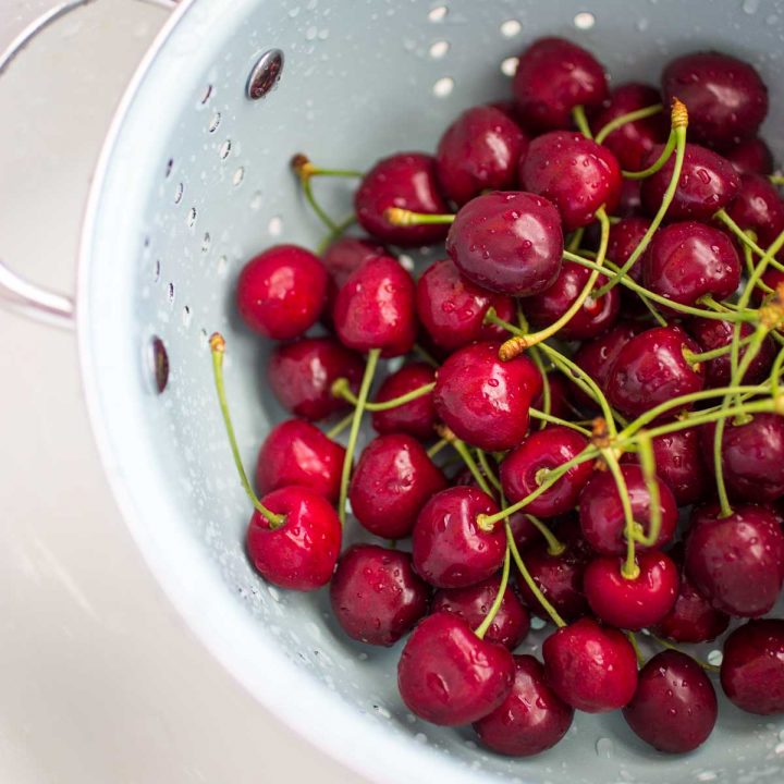 A blue strainer holds a pile of fresh cherries with stems for rinsing.