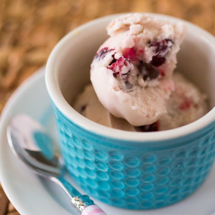 A blue bowl holds a scoop of homemade cherry chocolate chip ice cream which is pale pink with fresh cherries peeking out.