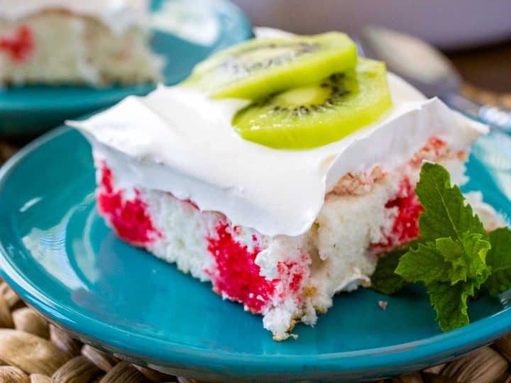 A square slice of strawberry kiwi cake shows the red Jello poke stripes, fluffy whipped topping, and has two thin slices of fresh kiwi on top for garnish.