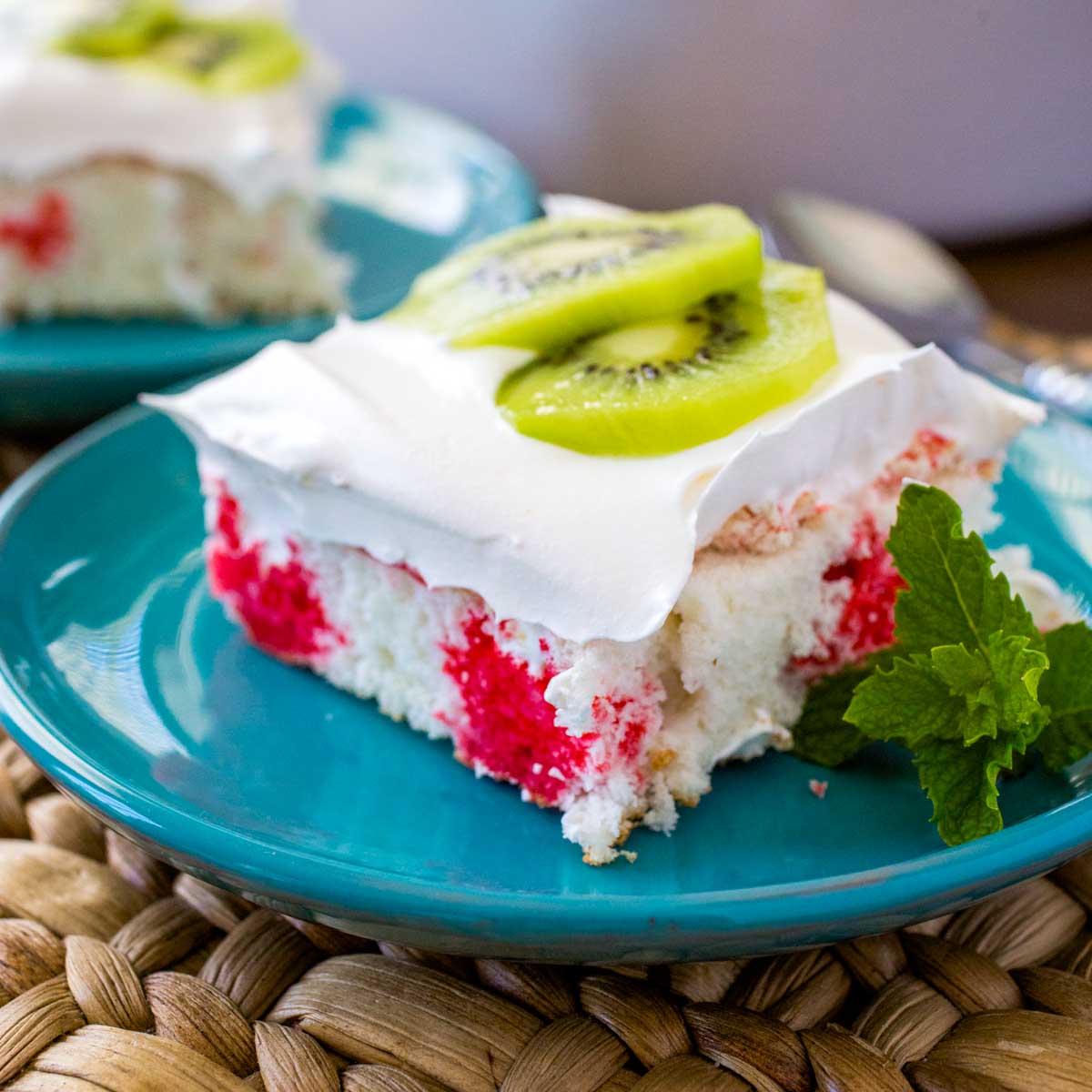A square slice of strawberry kiwi cake shows the red Jello poke stripes, fluffy whipped topping, and has two thin slices of fresh kiwi on top for garnish.