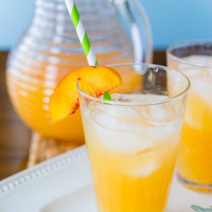 A glass of rosemary peach lemonade has lots of ice and a green striped straw. There's a fresh peach wedge on the glass for garnish. A pitcher of lemonade sits in the background.