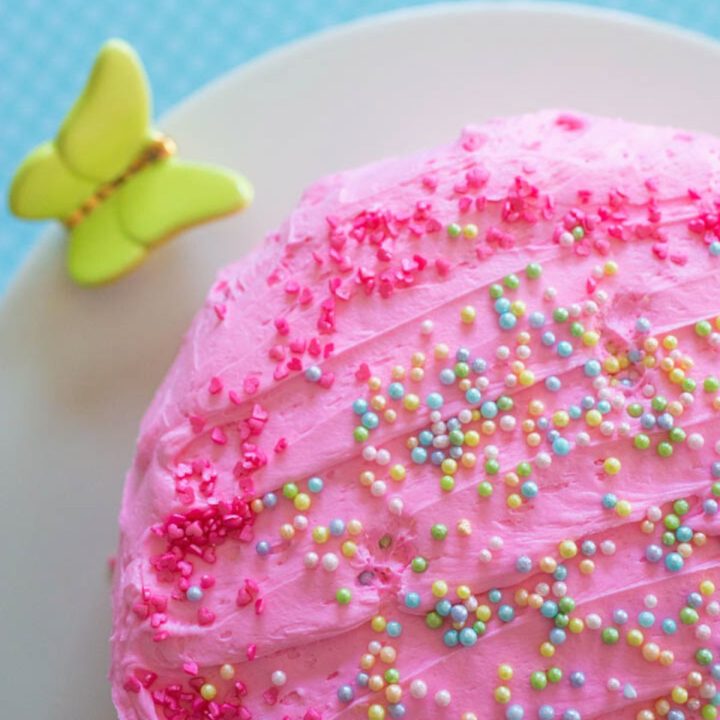 A pink birthday cake with sprinkles is on a cake stand with a butterfly.