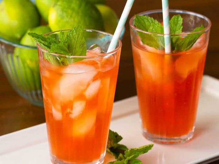 Two glasses of orange mojito mocktails sit on a platter with fresh mint. A bowl of limes sits in the background.