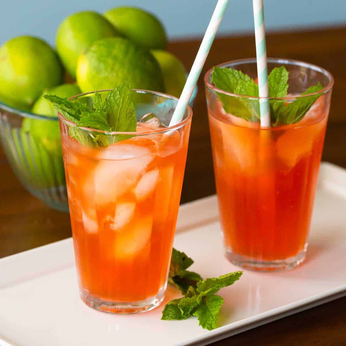 Two glasses of orange mojito mocktails sit on a platter with fresh mint. A bowl of limes sits in the background.