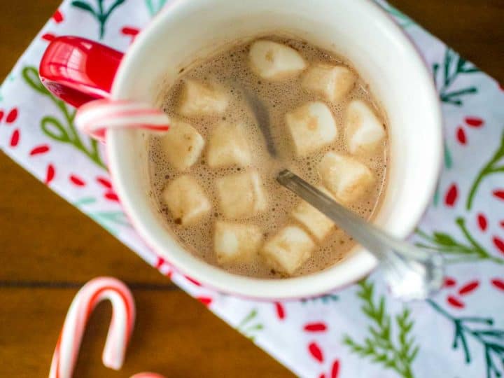 A red mug filled with hot cocoa and mini marshmallows has a candy cane hanging over the edge. Two mini candy canes are on the table and there's a festive holly napkin.