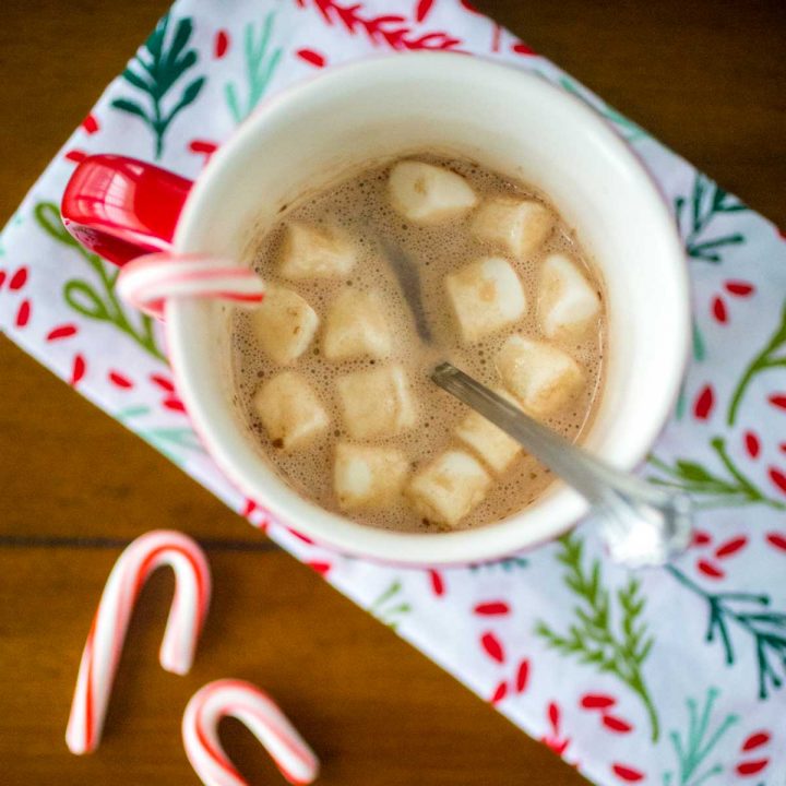 A red mug filled with hot cocoa and mini marshmallows has a candy cane hanging over the edge. Two mini candy canes are on the table and there's a festive holly napkin.
