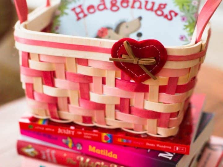 A pink and red basket with a felt heart has a Valentine's board book inside and sits on a stack of pink and red books.