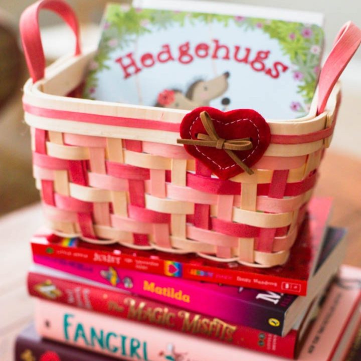 A pink and red basket with a felt heart has a Valentine's board book inside and sits on a stack of pink and red books.