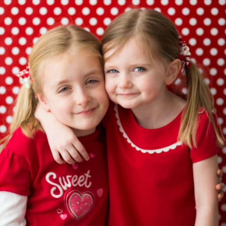 Two young girls wearing Valentine's Day shirts in front of a red and white polka dot background.