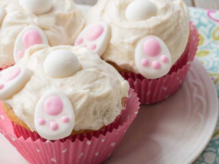 Easter bunny butt cupcakes in pink polka dot wrappers have white frosting and little decorative candies that look like bunny feet, a tail, and ears.