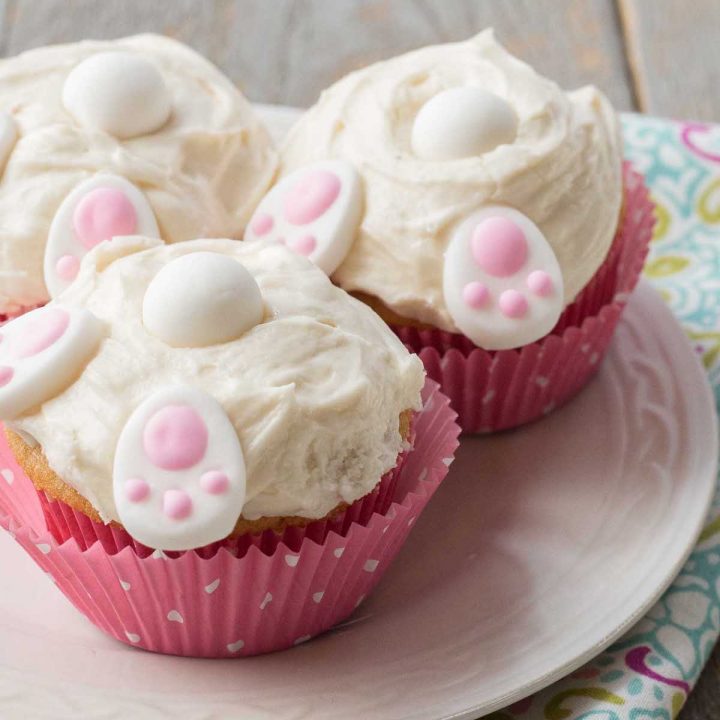 Easter bunny butt cupcakes in pink polka dot wrappers have white frosting and little decorative candies that look like bunny feet, a tail, and ears.