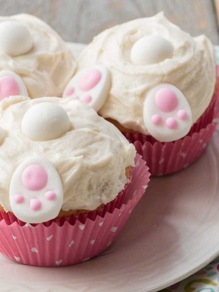 Easter bunny butt cupcakes in pink polka dot wrappers have white frosting and little decorative candies that look like bunny feet, a tail, and ears.