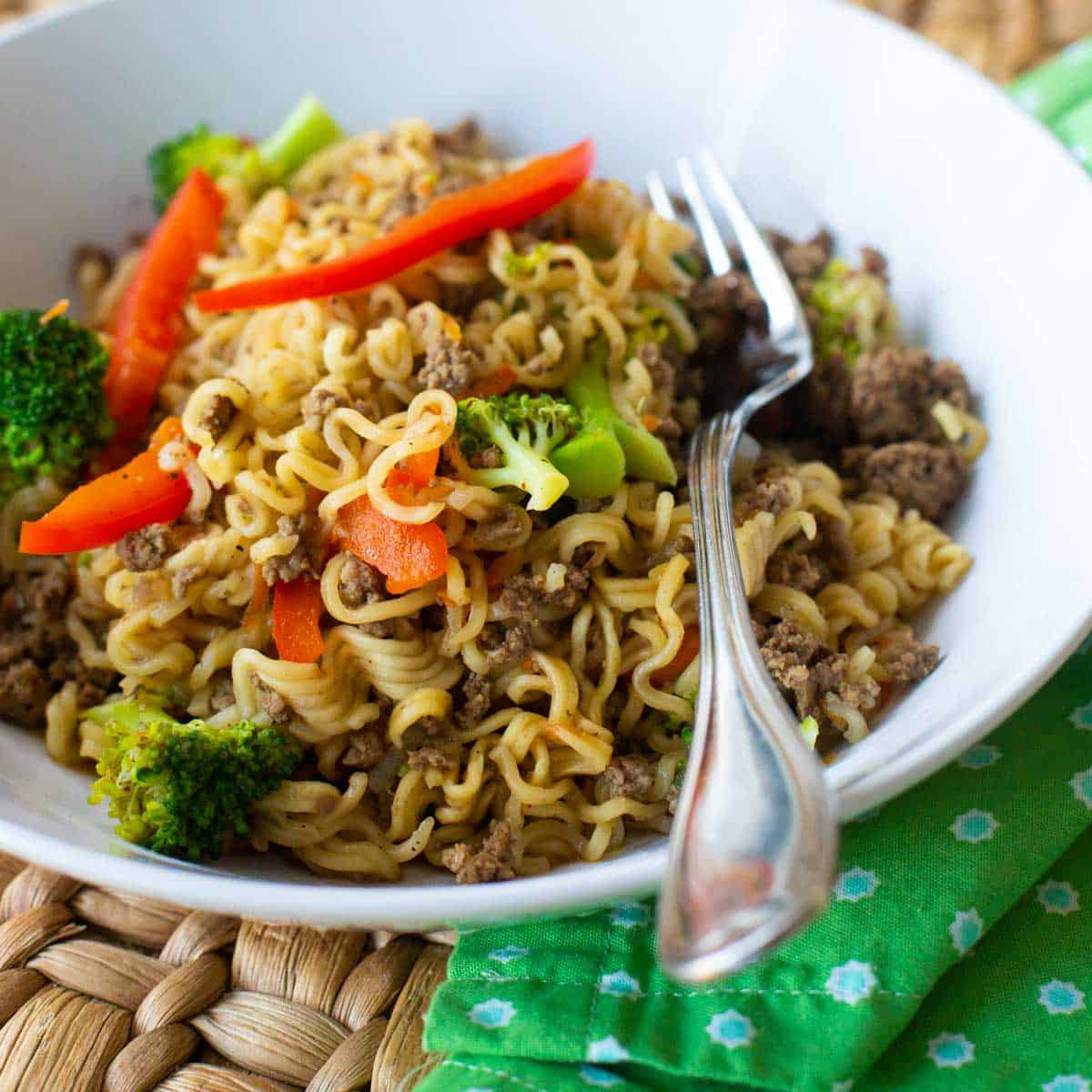 A white bowl of ramen noodles and ground beef with red peppers and broccoli.