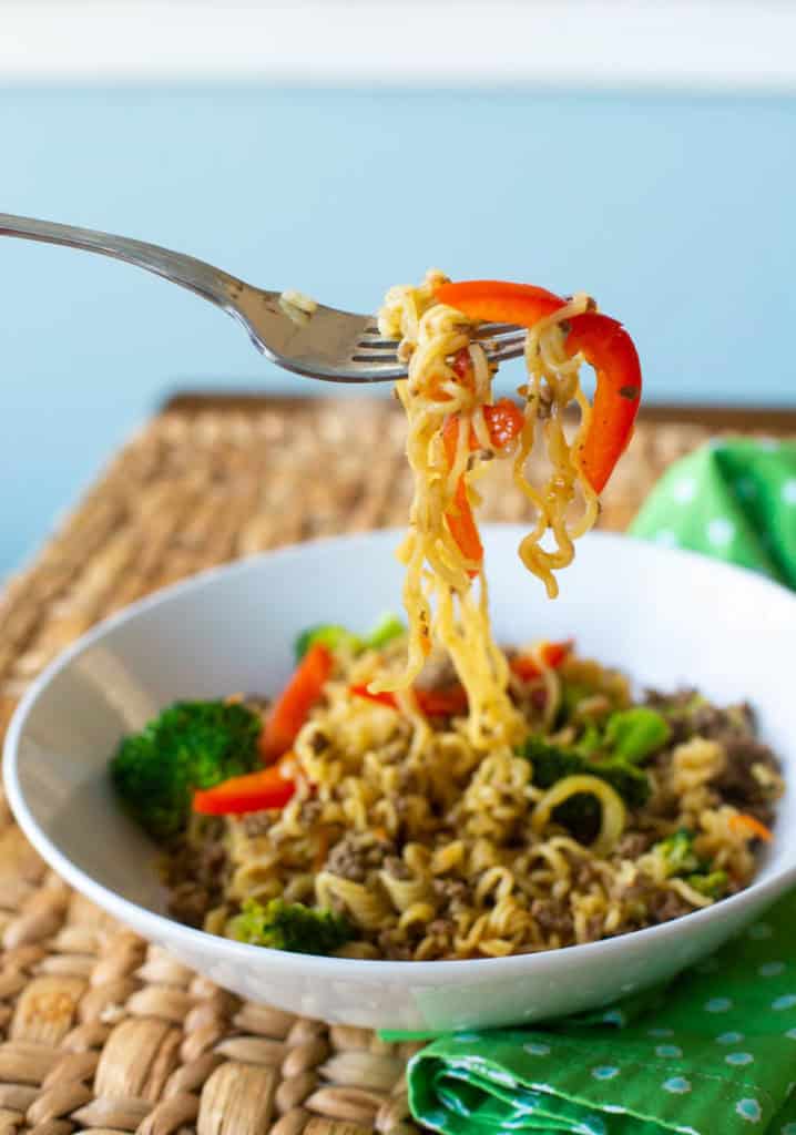 A white bowl filled with ramen and ground beef with fresh veggies. A fork is pulling up a bite.