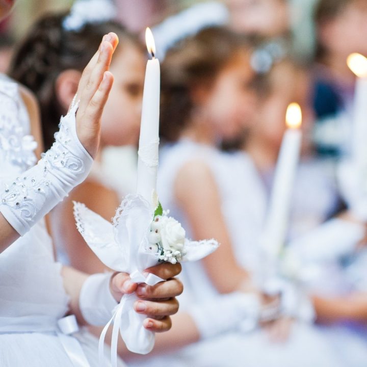 A group of young girls dressed in white dresses holding lit white candles for their First Communion Day.