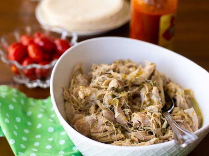 A bowl full of shredded chicken with a spoon. There's a bowl of red tomatoes, a green napkin, and a bottle of hot sauce sitting nearby.