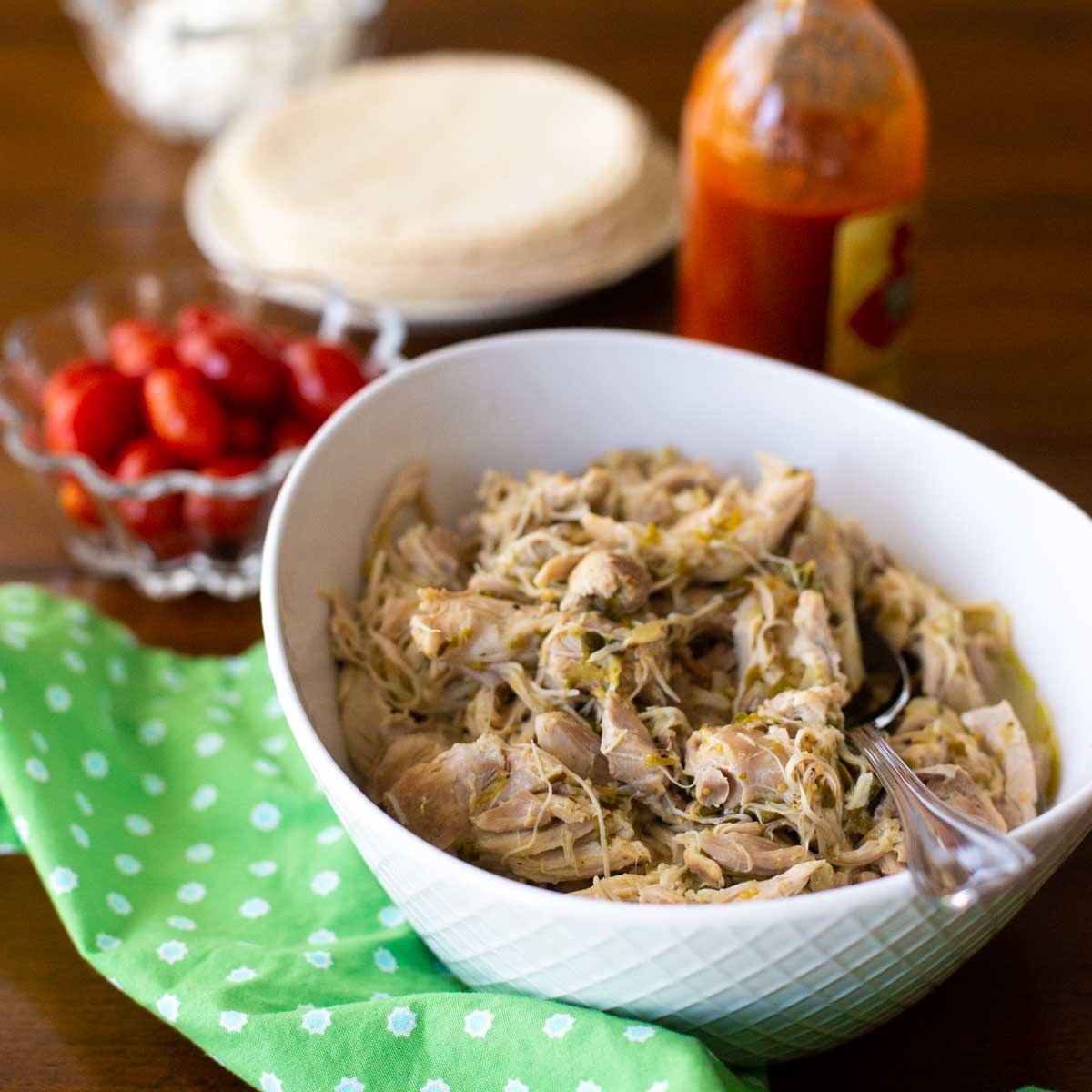 A bowl full of shredded chicken with a spoon. There's a bowl of red tomatoes, a green napkin, and a bottle of hot sauce sitting nearby.