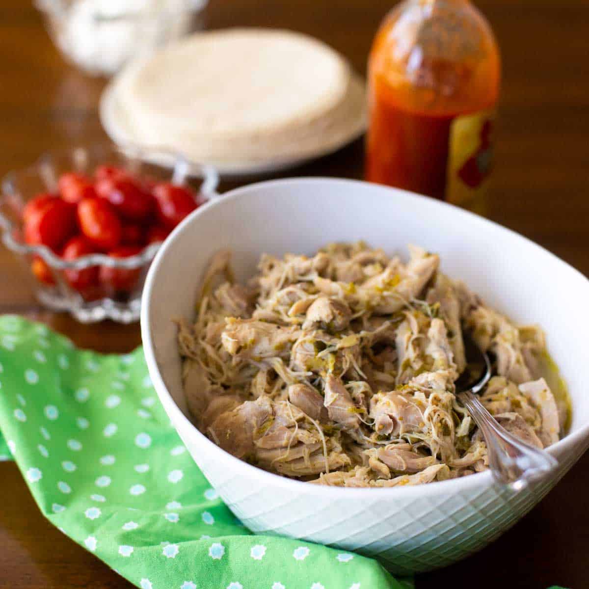 A bowl full of shredded chicken with a spoon. There's a bowl of red tomatoes, a green napkin, and a bottle of hot sauce sitting nearby.