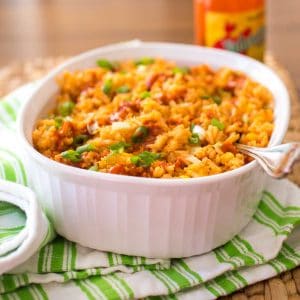 A white baking dish is filled with baked Mexican Rice. The tomato sauce base gave the rice a yellow-orange color. Hot sauce is drizzled over the top and fresh green onions are sprinkled for garnish.