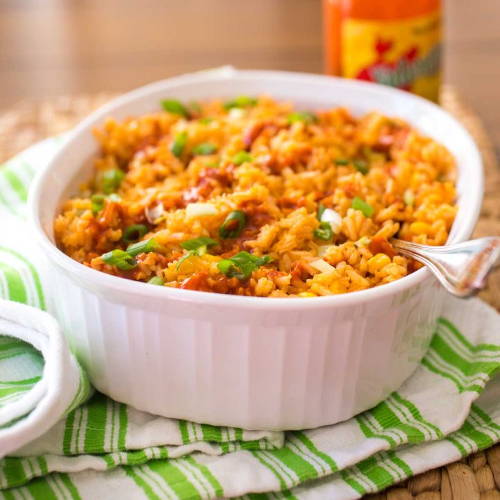 A white baking dish is filled with baked Mexican Rice. The tomato sauce base gave the rice a yellow-orange color. Hot sauce is drizzled over the top and fresh green onions are sprinkled for garnish.