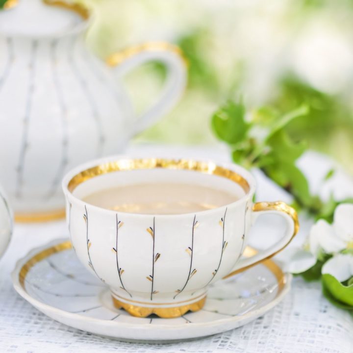 A gilded tea cup filled with milky tea sits on a table with pretty green and white flowers.
