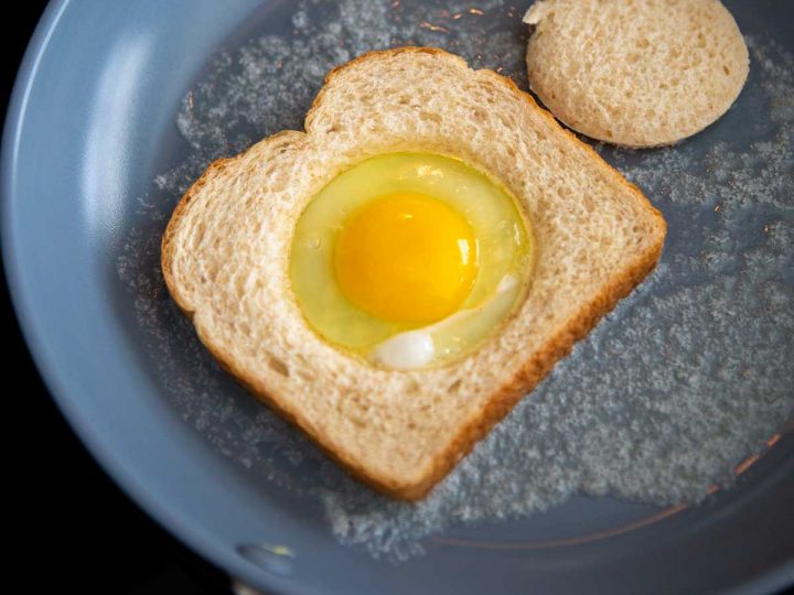 A fry pan with melted butter and a slice of bread with the center hole missing. An egg has been cracked into the hole.
