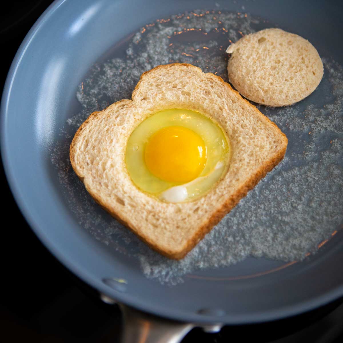 A fry pan with melted butter and a slice of bread with the center hole missing. An egg has been cracked into the hole.