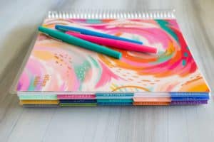 An Erin Condren planner is on a table with 3 pens on top.