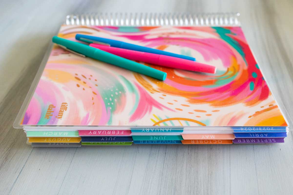 An Erin Condren planner is on a table with 3 pens on top.