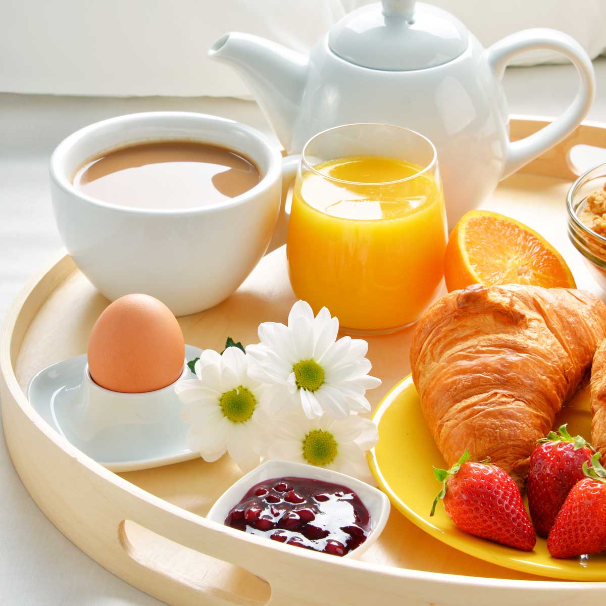 A round tray holds a white tea pot, a coffee cup with milky coffee, 3 fresh daisies, a brown egg, and a yellow plate with a croissant and fresh strawberries with a tiny cup of jam.