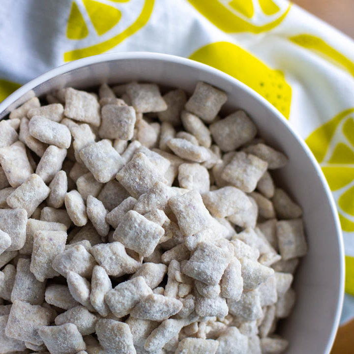 A bowl of powdered sugar covered Chex cereal has been made into muddy buddies. A lemon printed napkin is on the side.