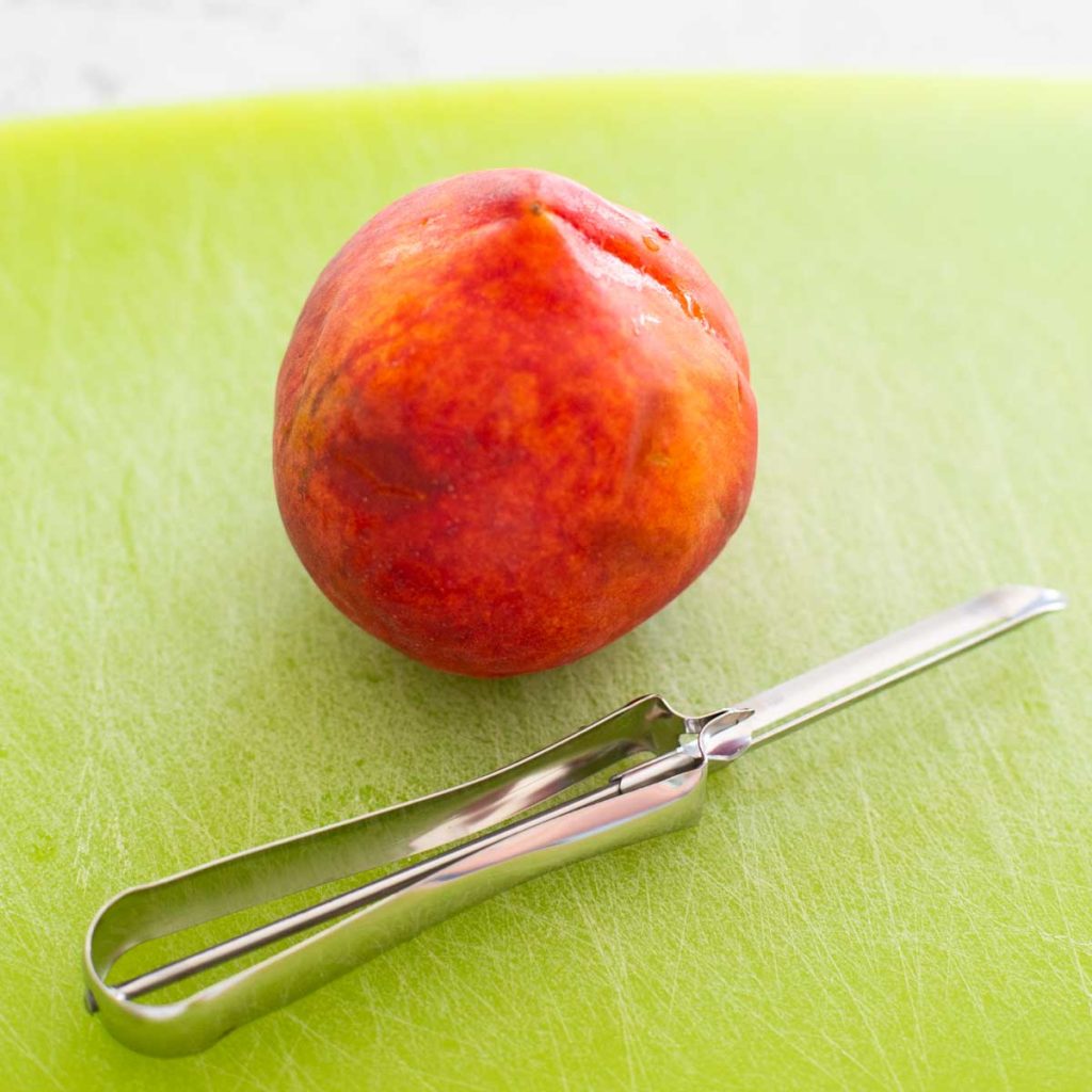 A fresh peach next to a peeler on a cutting board.