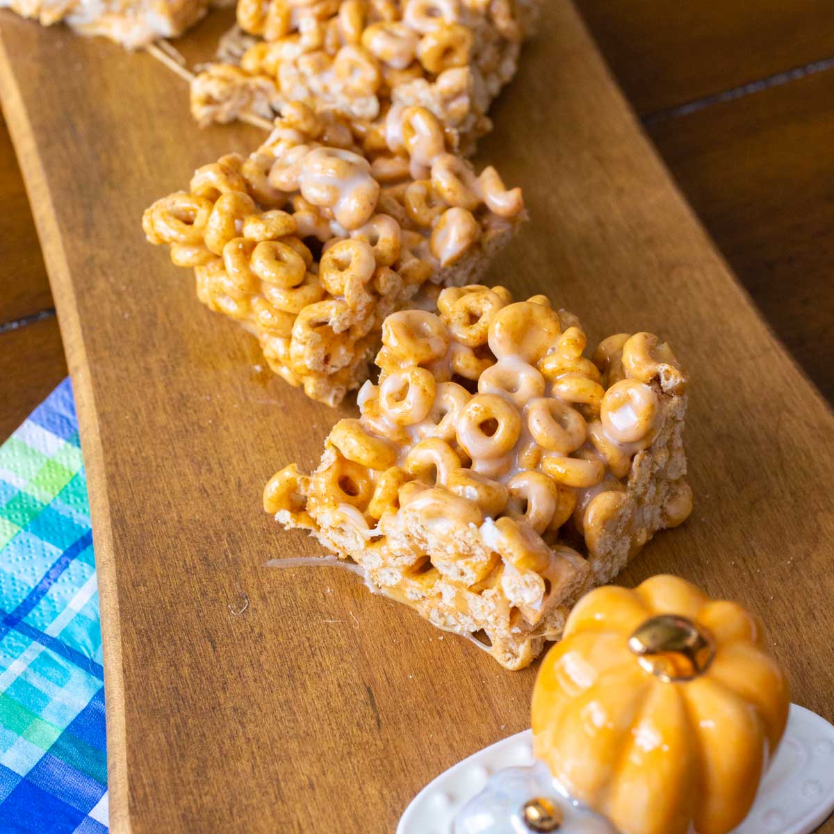 A wooden platter has 3 squares of pumpkin spice cheerio bars with a ceramic pumpkin decoration.