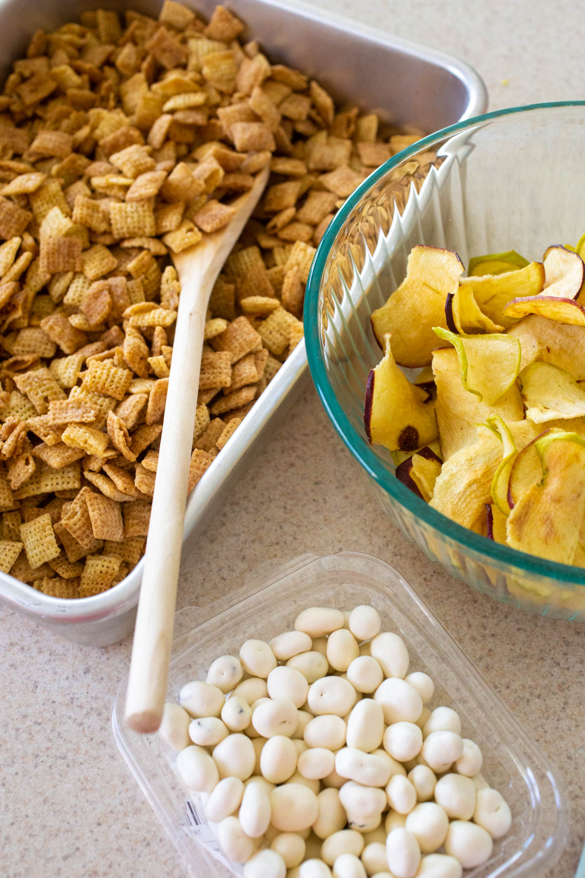 The ingredients are in separate bowls about to be stirred together. The Chex cereal is in a pan, apple chips in a mixing bowl, and yogurt covered raisins in a small package.