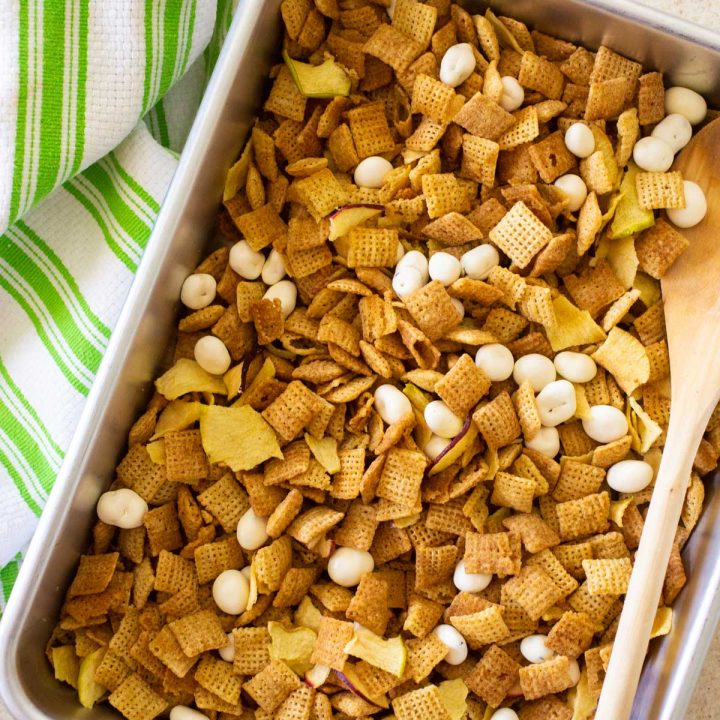 A pan of apple pie chex mix has yogurt covered raisins mixed in with a wooden spoon next to a green striped towel.