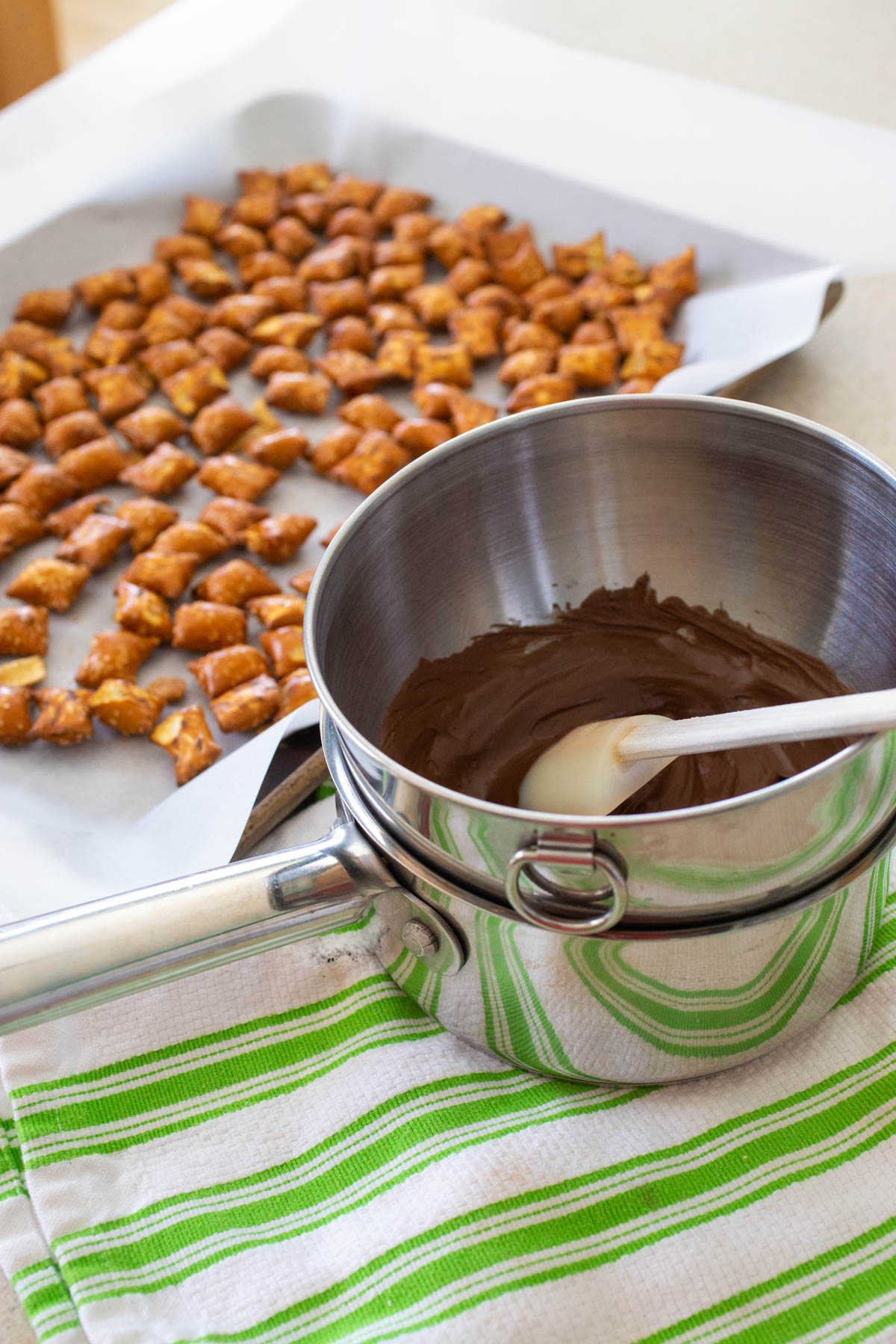 The chocolate chips have been melted in a double boiler made out of a mixing bowl and a sauce pan. The pretzels are ready to be drizzled with chocolate on a baking pan in the background.