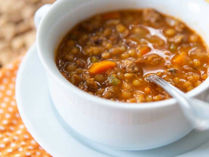 A bowl of sausage lentil soup sits on an orange napkin with a spoon ready for eating.