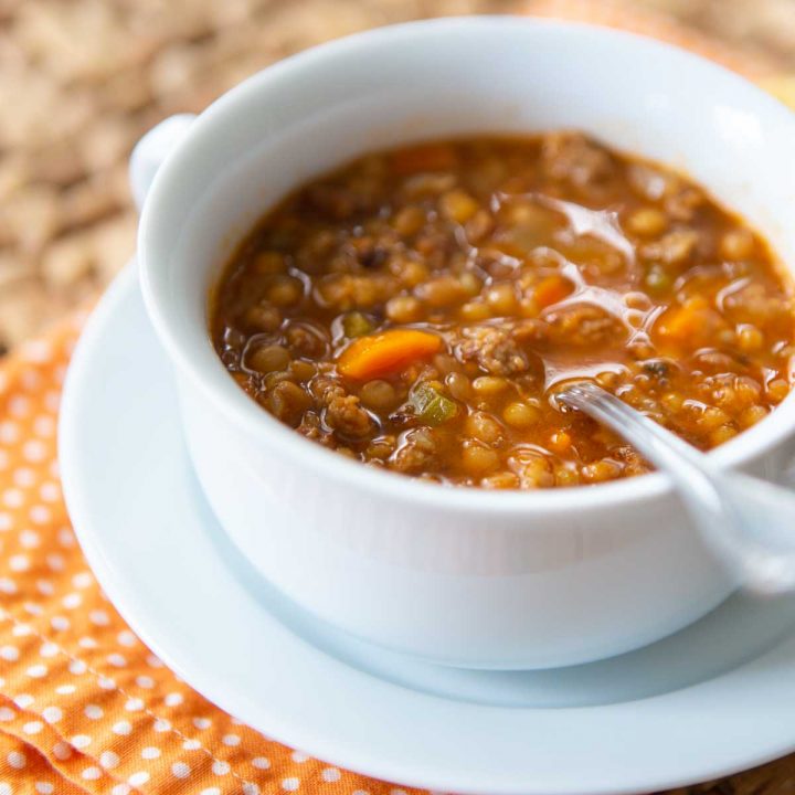 A bowl of sausage lentil soup sits on an orange napkin with a spoon ready for eating.