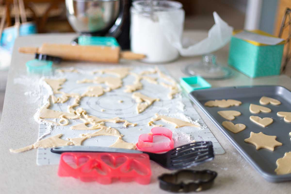 A kitchen counter is filled with messy tools from a baking day for sugar cookies. Dough, cutters, a mixing bowl and rolling pin and a messy spatula.