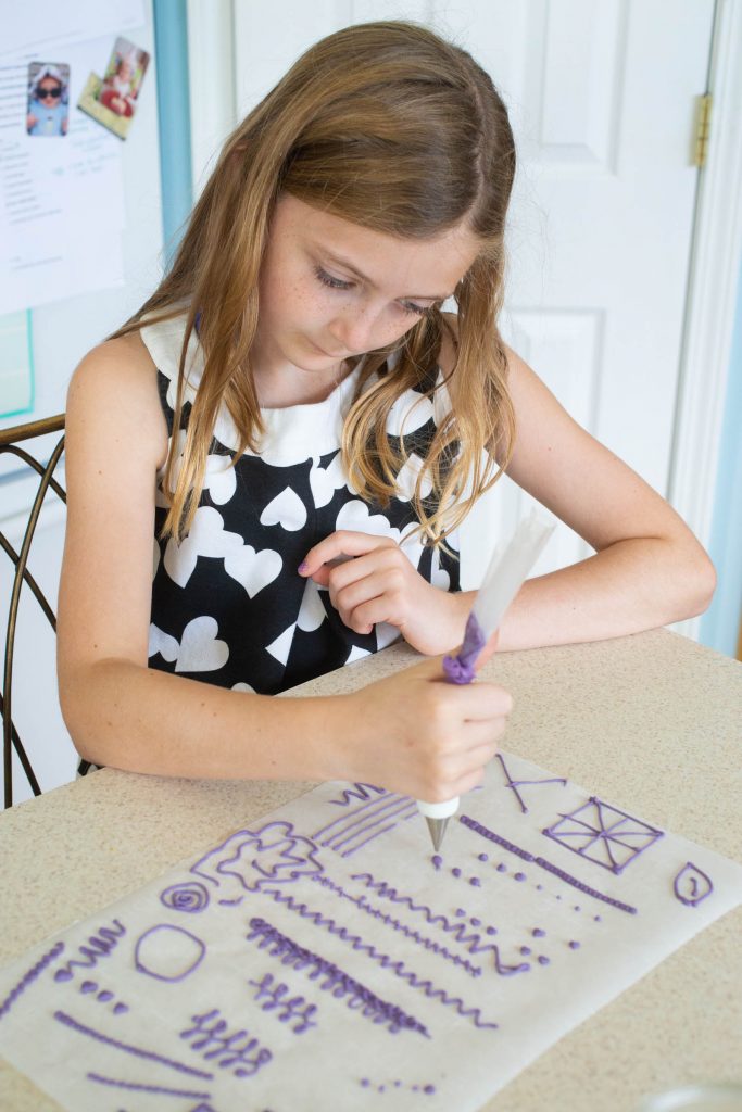 A young girl practices piping with purple icing on a piece of parchment paper.