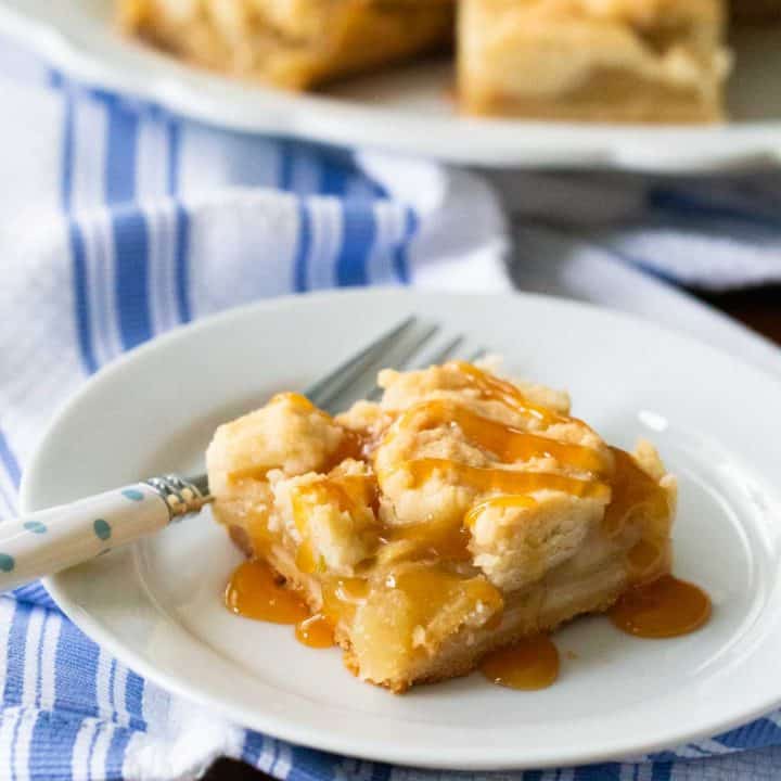 A square of apple crumble bars is served on a white plate with a drizzle of caramel sauce over the top.