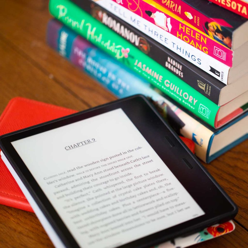 A Kindle reader sits on a table next to a stack of books.