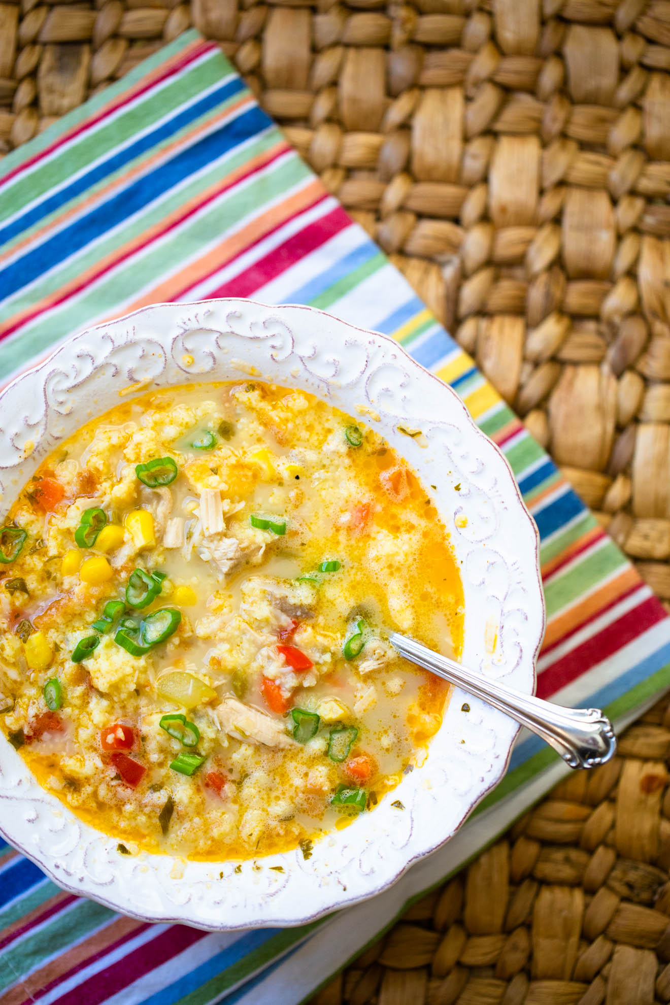 A bowl of chicken soup has bright yellow corn, red peppers, and green onions over the top.