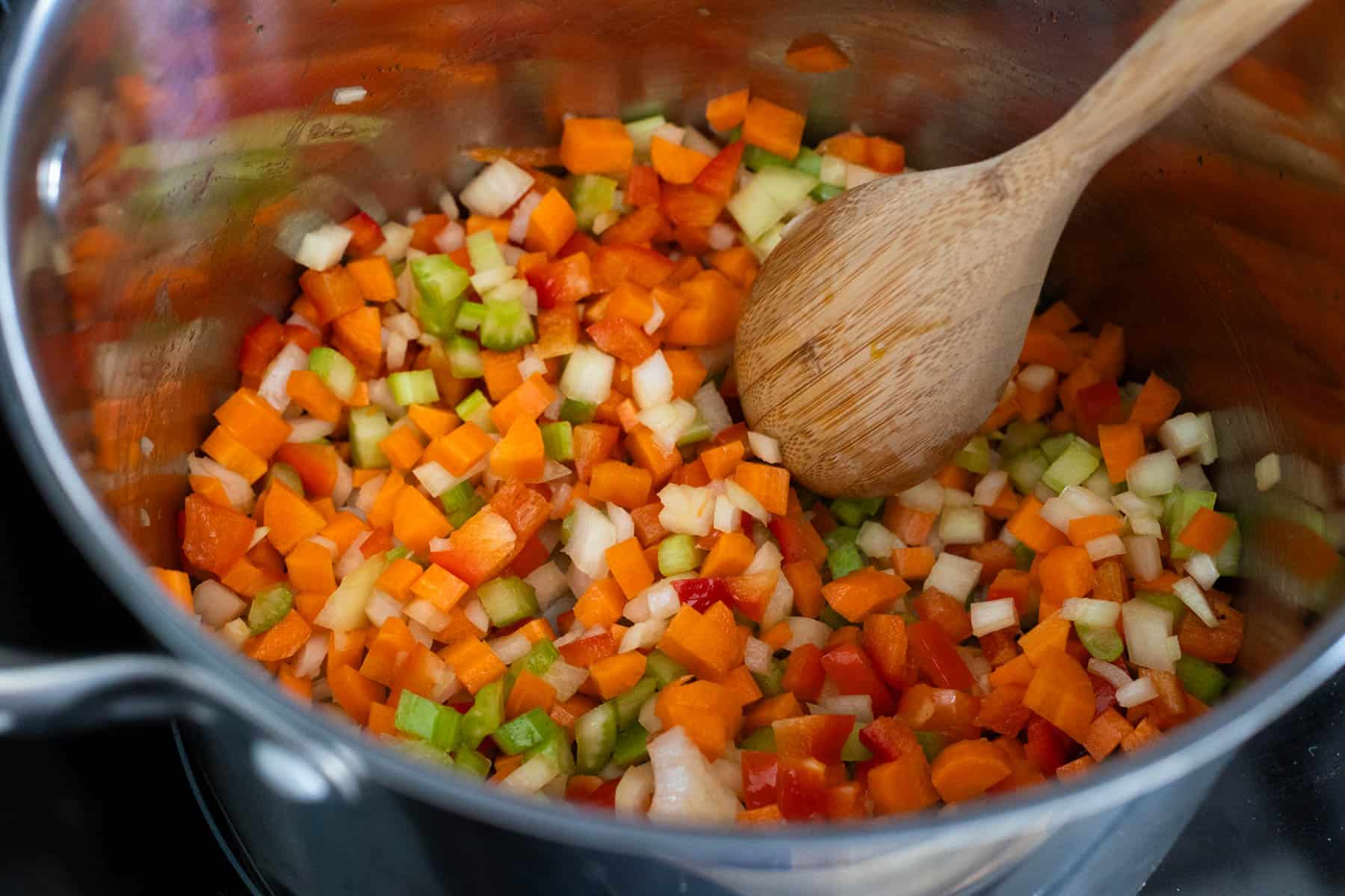 The soup pot has all the veggies in it, a wooden spoon is stirring them. 