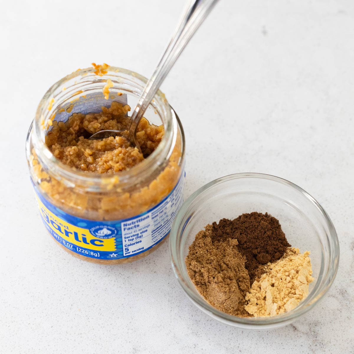 The jar of minced garlic has a spoon in it and sits next to a small prep cup of dried seasonings.