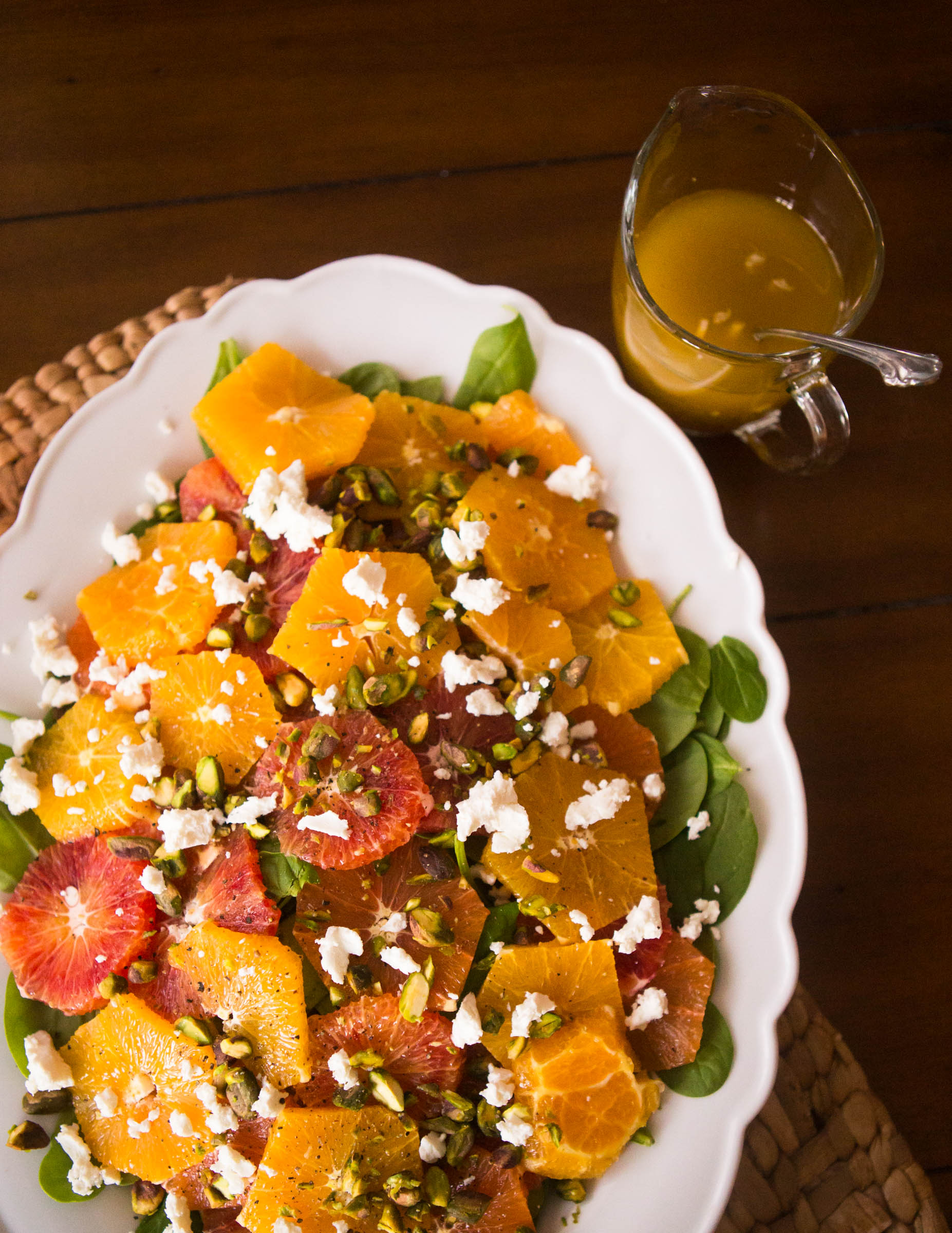 Sliced citrus salad features fresh oranges, baby spinach, and pistachios in layers on a ruffled white platter next to a serving pitcher of homemade dressing.